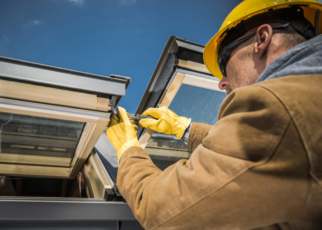 Caucasian Worker Wearing Safety Helmet and Protective Gloves Carrying Out Repair Works on Roof Skylight Windows Using His Screwdriver.